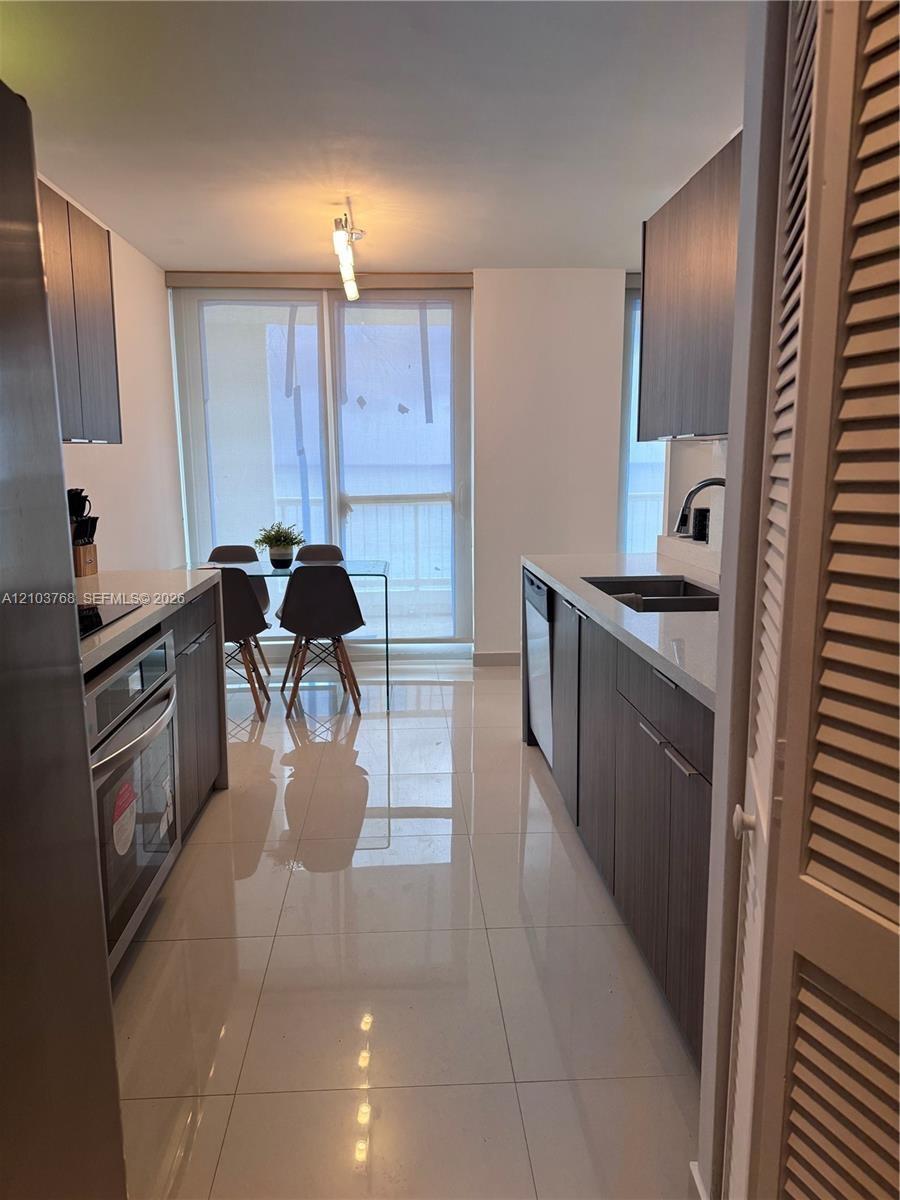 Galley-style kitchen featuring flat-panel cabinetry, light-toned stone countertops, polished tile flooring, and stainless steel built-in appliances