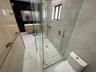 Walk-in glass-enclosed shower featuring gold-toned hardware, wall-mounted vanity with dual sinks, large-format tile flooring, and contrasting black an