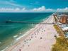 Aerial perspective of coastal terrain featuring an expansive sandy beach, turquoise ocean waters, and a prominent public fishing pier