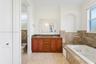 Dual-sink vanity with wood-finish cabinetry, natural stone countertop, recessed wall niche, tiled soaking tub, and stone-finish flooring