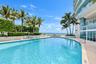 Curved swimming pool with concrete decking, surrounding palm trees, low-profile masonry wall, and glass-paneled building exterior
