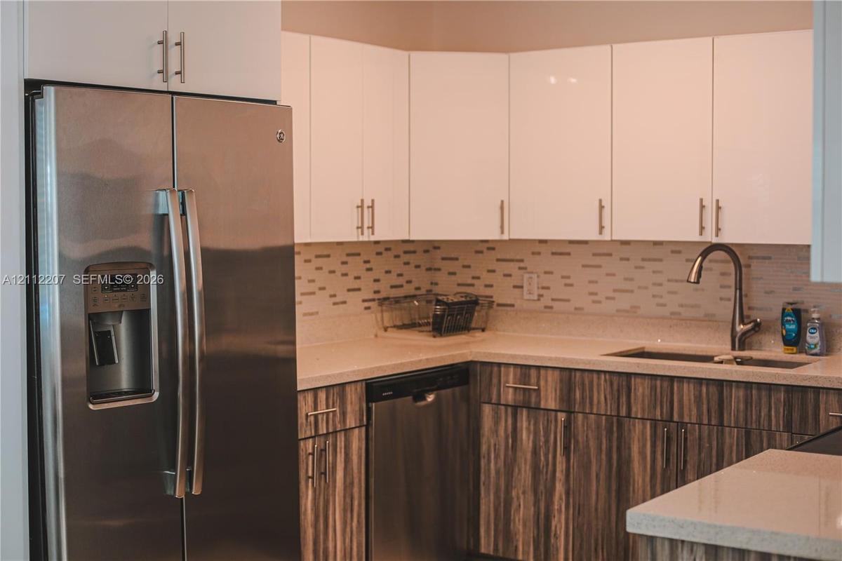 Kitchen featuring white upper cabinetry, wood-finish lower cabinetry, stone-surface countertops, a stainless steel refrigerator and dishwasher, and a 