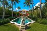 Custom in-ground swimming pool featuring a shaped stone-finished coping, central decorative sculpture, terracotta tile roof architecture, and mature p