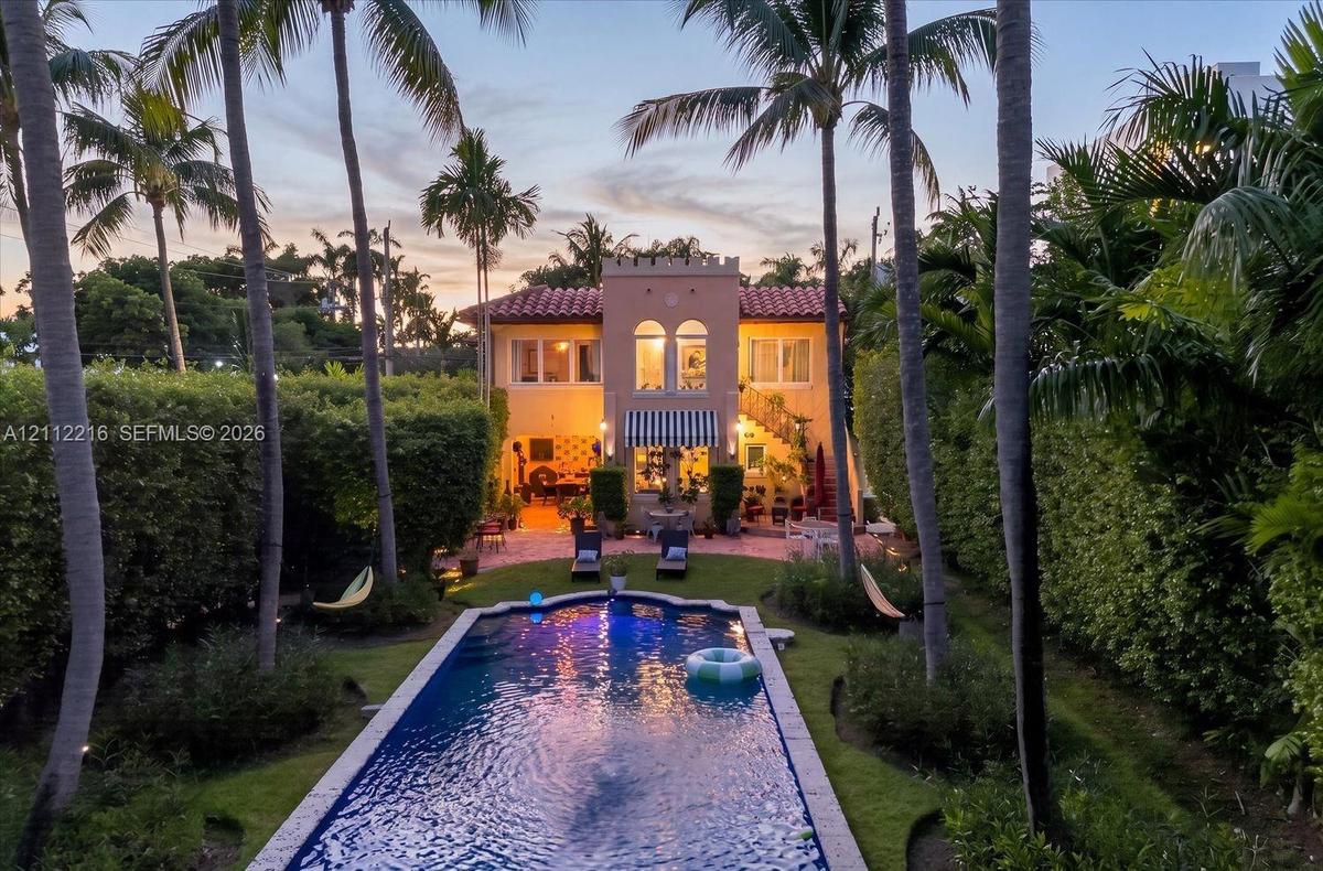Spanish Colonial architecture featuring a crenellated tower, red tile roof, rectangular swimming pool, brick-paved patio, and mature palm trees