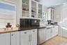 Kitchen featuring white cabinetry with glass-front uppers, dark stone countertops, stainless steel dishwasher, farmhouse-style sink with wall-mounted 