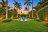 Rectangular swimming pool with stone coping, surrounded by a manicured lawn, established palm trees, and dense perimeter hedging