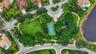 Aerial perspective of a community recreation area featuring a basketball court, playground, expansive green space, mature trees, and a bordering water