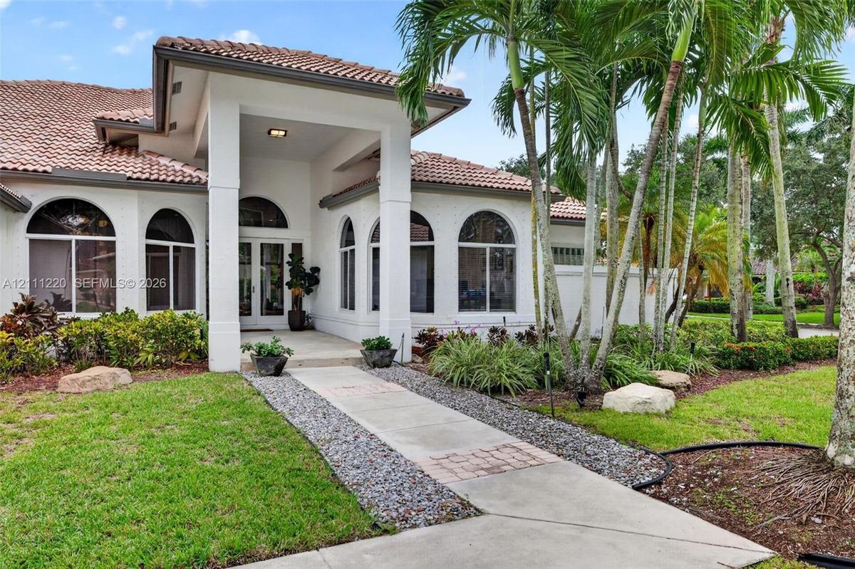 Mediterranean-style exterior featuring a covered entryway, stucco facade, arched windows, concrete walkway, and mature palm trees