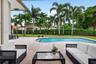 Covered patio featuring a concrete deck, curvilinear swimming pool, manicured lawn, and perimeter hedging with mature palm trees