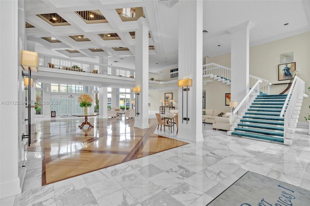 Grand foyer featuring a coffered ceiling, polished stone flooring with wood-finish inlays, structural columns, and an open staircase with a custom rai