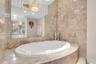 Oval soaking tub featuring a decorative basin, surrounded by large-format natural stone tiling with an expansive wall-to-wall mirror, crystal-style ch