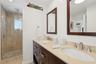 Double vanity with stone countertop, dark wood-finish cabinetry, frameless glass shower enclosure, stone-tiled shower wall, and integrated shower rain