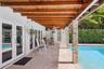 Covered patio featuring a wood-beam pergola, stone-clad support columns, natural stone flooring, and built-in glass block bar, complemented by a swimm