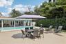 Spacious paved patio featuring an in-ground swimming pool, stone-accented exterior wall, and dense mature perimeter landscaping