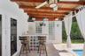 Covered patio featuring an exposed wood-beam ceiling, ceiling fan with integrated lighting, tiled bar with glass-block base, and stone-finish flooring