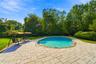 Inground swimming pool with metal ladder, surrounded by an expansive stone paver patio, set within a level lawn bordered by mature trees and shrubs