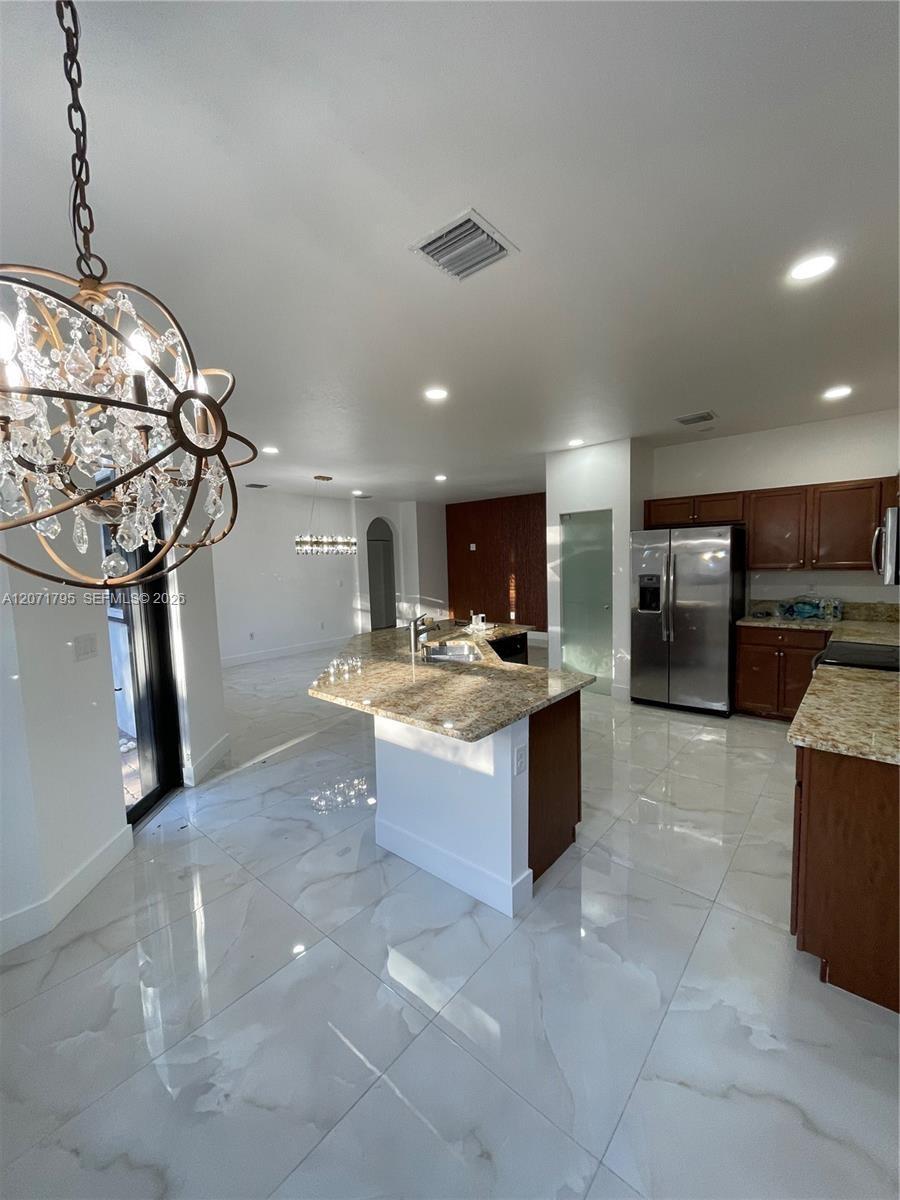 Kitchen featuring stone-finish countertops, dark wood cabinetry, large-format tile flooring, a center island with an integrated sink, and an ornamenta