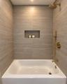 Bathtub with integrated shower featuring large-format tile surround, recessed wall niche, and brushed gold finish fixtures including a rainfall shower