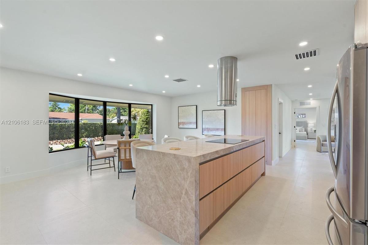 Kitchen featuring a waterfall-edge center island, stone countertops, flat-panel light wood cabinetry, a cylindrical stainless steel range hood, and la