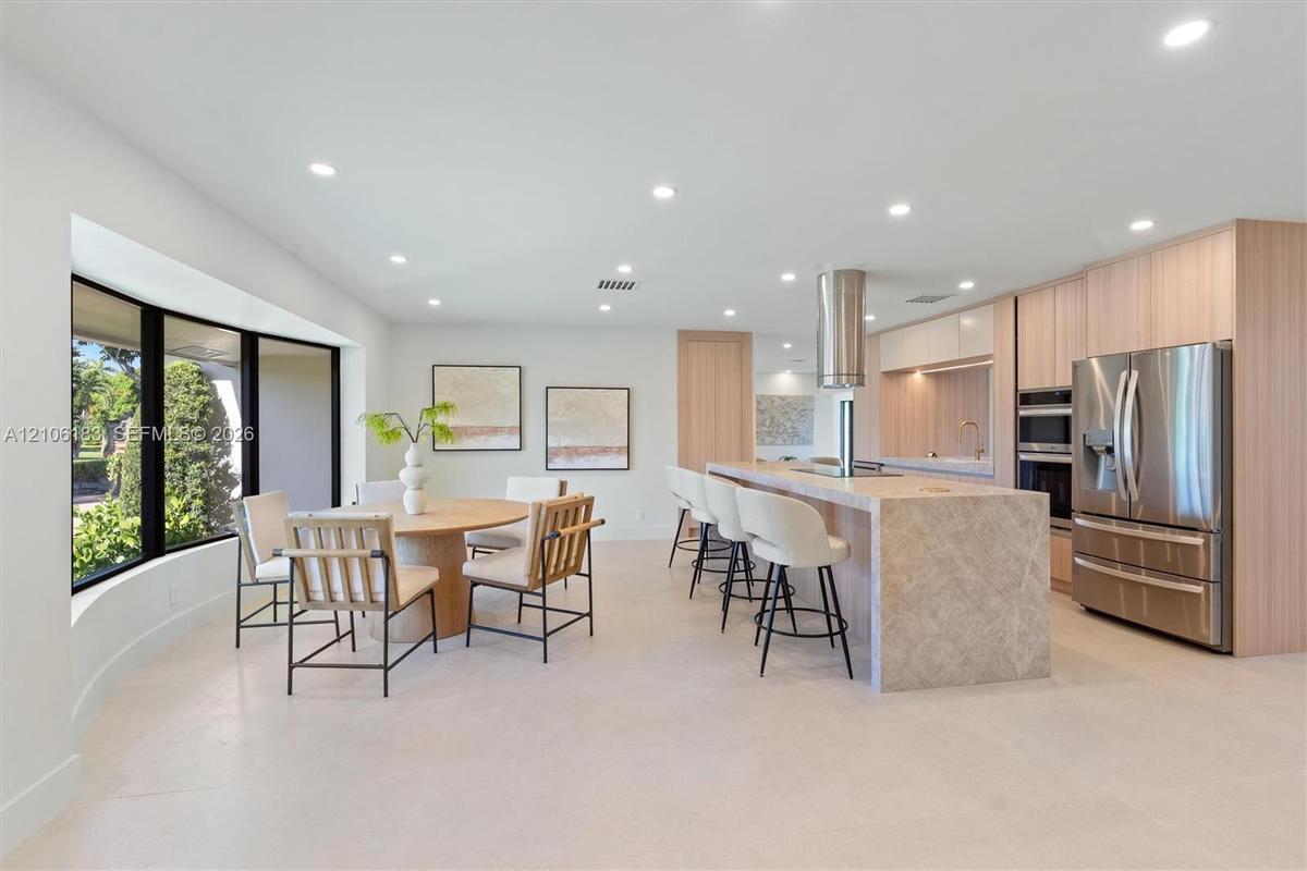 Modern kitchen featuring light wood-finish cabinetry, a large stone-surfaced island with a cylindrical exhaust hood, stainless steel appliances, reces