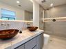 Bathroom vanity featuring a stone countertop, dual glass vessel sinks, wall-mounted gold-tone faucets, and a walk-in shower with floor-to-ceiling tile