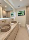 Floating vanity with dual fluted vessel sinks, stone-finish countertop, wall-mounted brass faucets, back-lit vanity mirror, and large-format tile wall