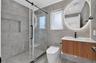 Bathroom featuring large-format gray wall and floor tile, walk-in glass shower with a built-in recessed shelf, wall-mounted wood-finish vanity, and il