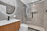 Floating wood-finish vanity with a white rectangular basin, circular LED-backlit mirror, large-format grey tile walling, and glass-enclosed shower wit