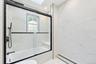 Full-height stone-finish wall tile, bathtub with integrated bench seating, black-framed glass sliding shower enclosure, matte black rainfall shower he