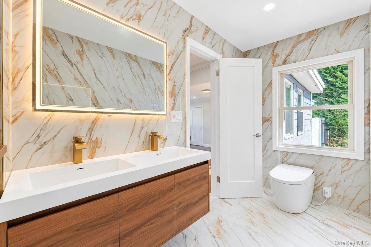 Floating dual-sink vanity with wood-finish cabinetry, quartz countertop, gold-tone fixtures, backlit rectangular mirror, and floor-to-ceiling stone-pa
