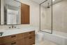 Wood-finish vanity with a rectangular sink, stone-composite countertop, matte black hardware, rainfall shower head, and glass partition