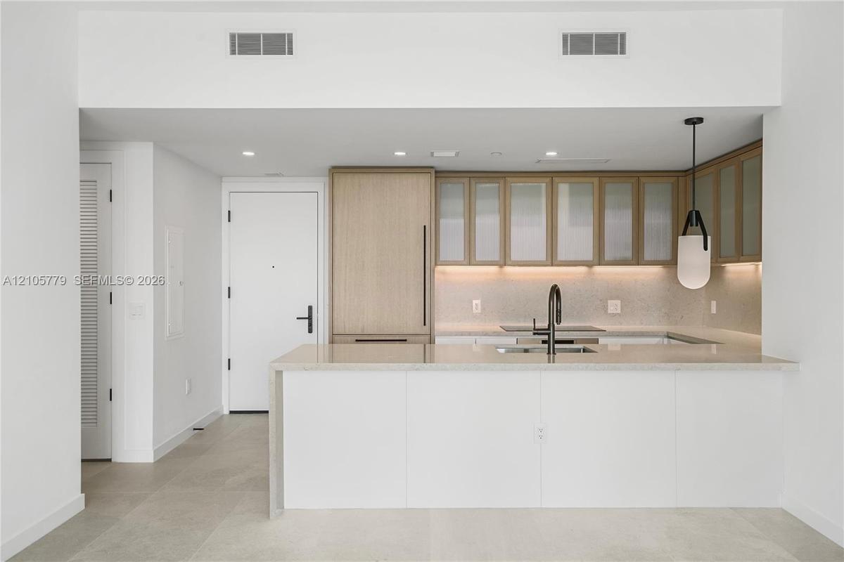 Kitchen featuring light wood-finish cabinetry with frosted glass inserts, a white quartz waterfall-edge island, integrated refrigerator, matte black g