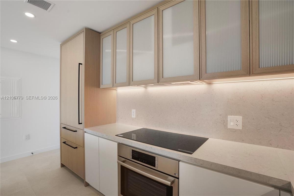 Custom cabinetry featuring light wood finish and reeded glass upper cabinet doors, integrated refrigeration, stone-finish countertops, and an inductio