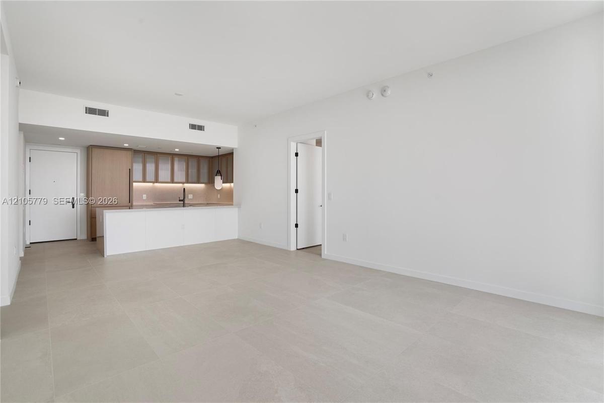 Open-concept interior with large-format neutral tile flooring, minimalist kitchen featuring light wood-tone cabinetry, stone-surface breakfast bar, an