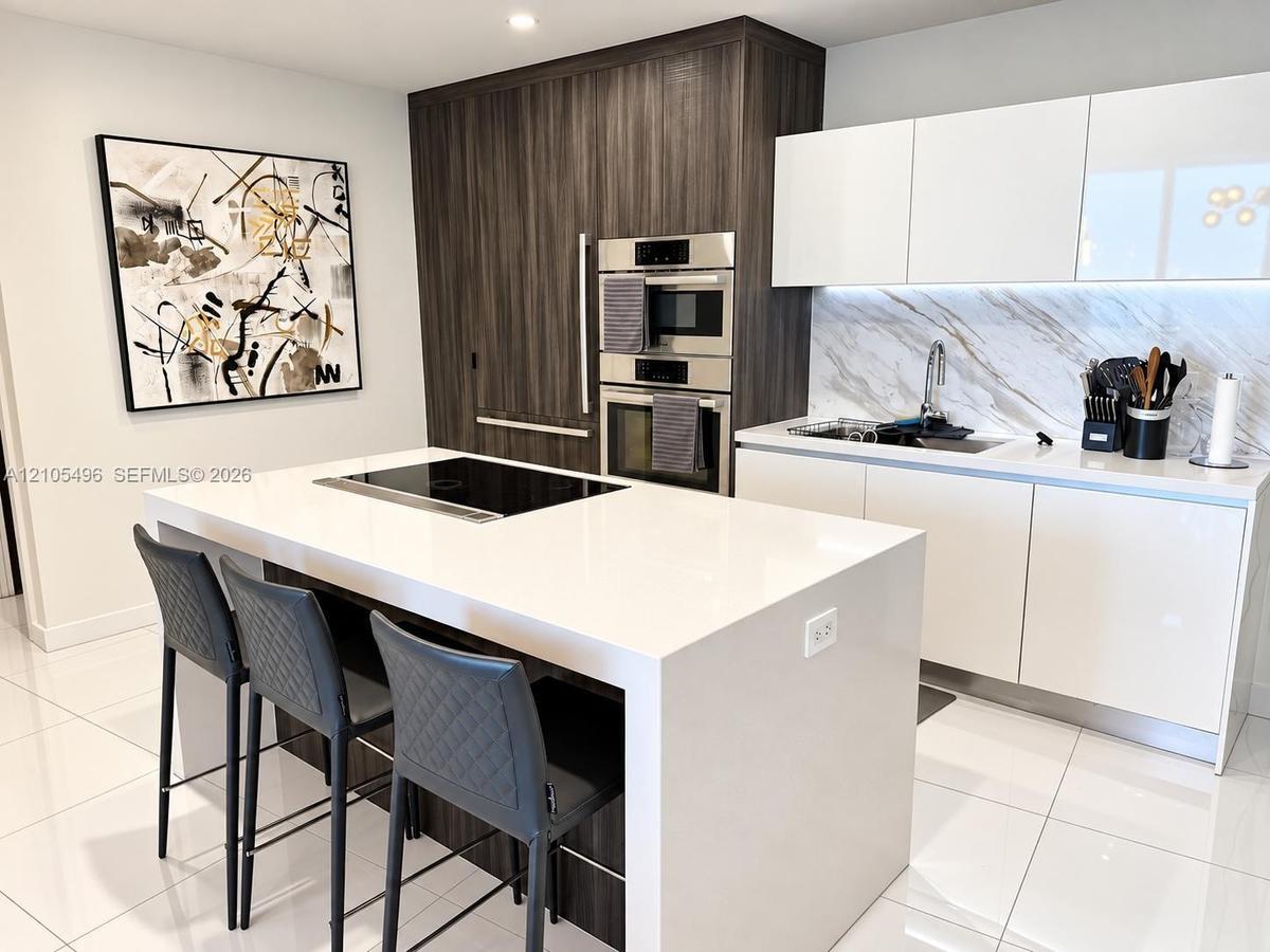 Kitchen featuring a waterfall-edge island with an integrated cooktop, flat-panel cabinetry, a stone-pattern backsplash, and stainless steel built-in a