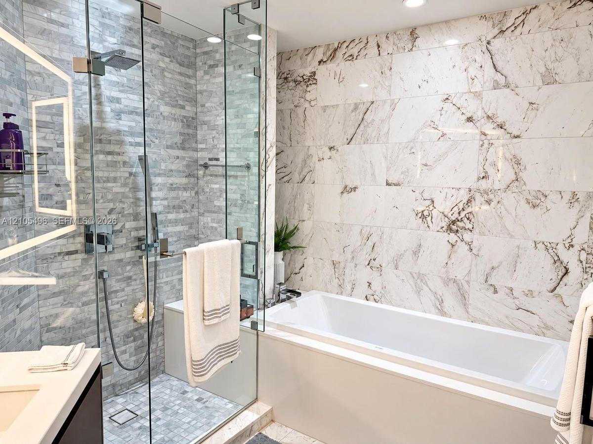 Walk-in glass-enclosed shower with stacked stone tiling, built-in soaking tub featuring a decorative stone-patterned wall surround, and a backlit vani