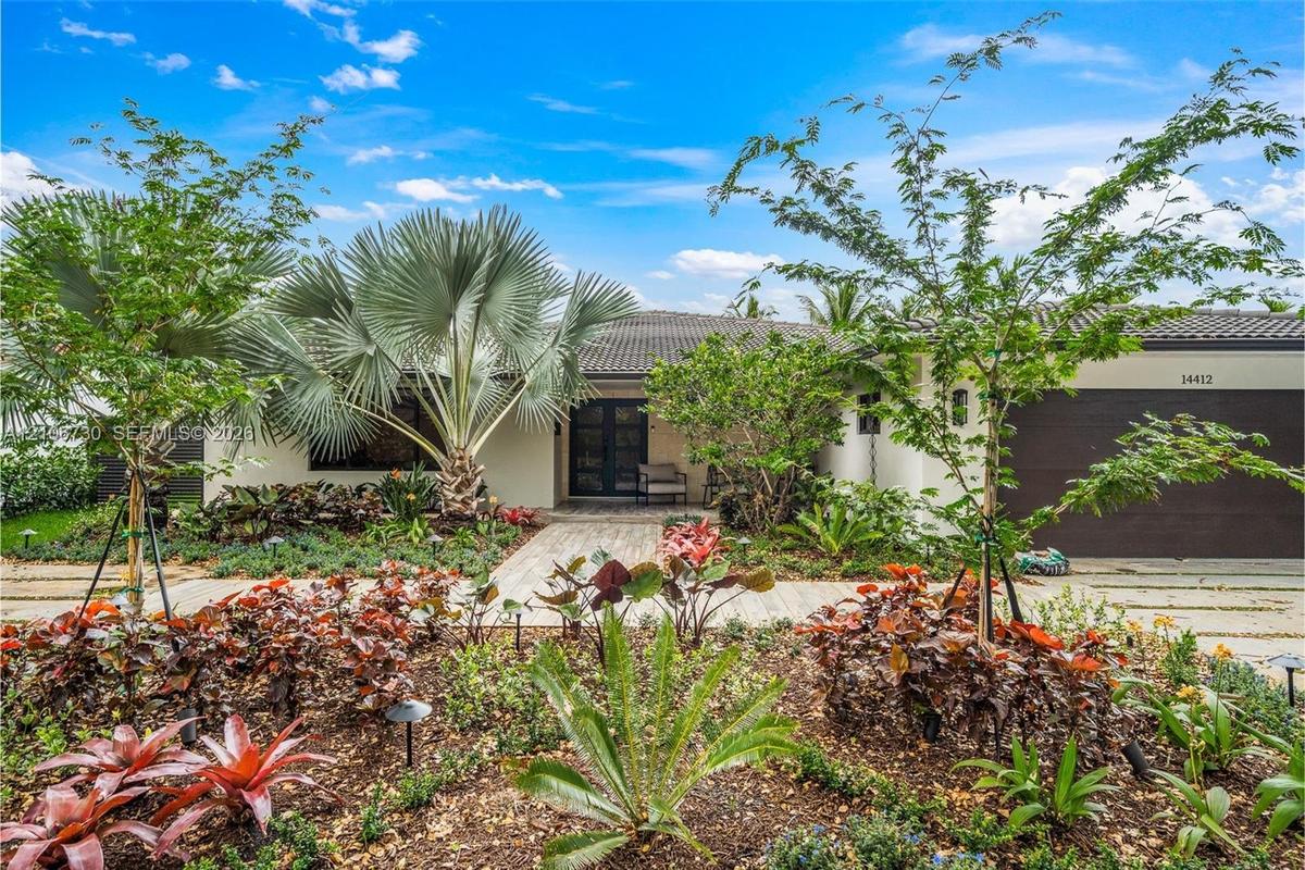 Single-story exterior featuring a tile roof, paved walkway, integrated multi-car garage, and professional landscaping with mature palms and diverse or