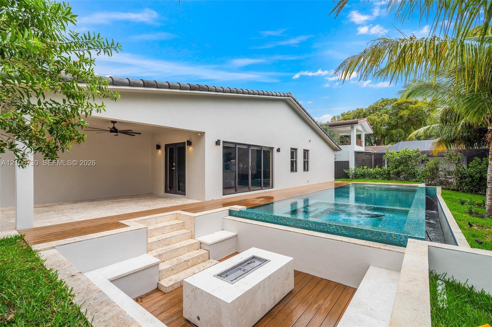 Covered outdoor patio with stone flooring, custom zero-edge pool, integrated stone fire feature, and tiered stone steps leading to a wood-finish deck
