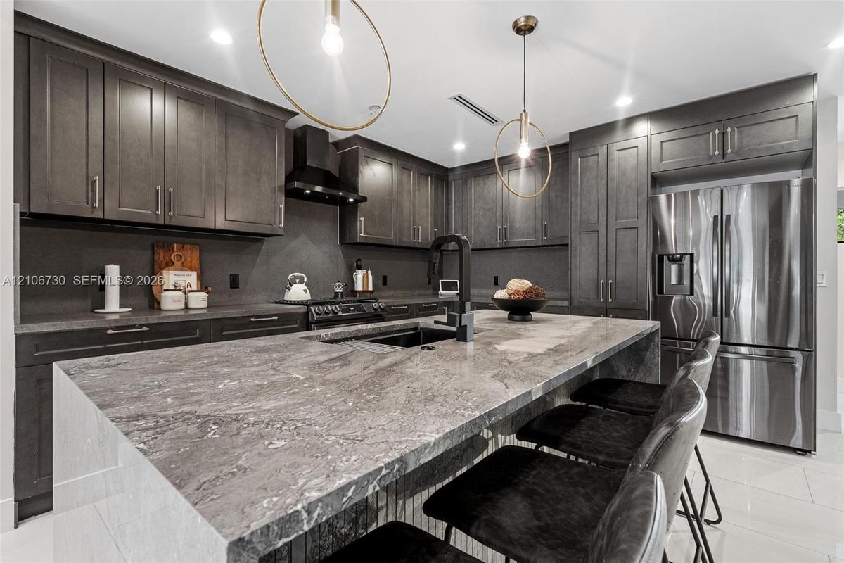 Kitchen featuring dark custom cabinetry, a stone-topped waterfall island, stainless steel appliances, a black range hood, and modern pendant lighting
