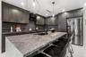 Kitchen featuring dark custom cabinetry, a stone-topped waterfall island, stainless steel appliances, a black range hood, and modern pendant lighting