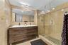 Dual-sink vanity with wood-finish cabinetry, full-width wall mirror, glass-enclosed shower, and stone tile surround