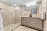 Bathroom featuring a walk-in glass-enclosed shower with a rainfall showerhead, wood-finish vanity with dual integrated sinks, and neutral stone-style 