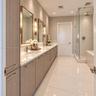 Dual-sink vanity with light-tone wood cabinetry, stone-finish countertops, brass-finish wall-mounted lighting, and a glass-enclosed walk-in shower