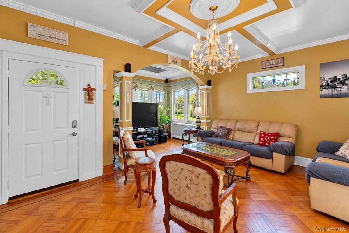 Living area featuring parquet wood flooring, coffered ceiling with a crystal chandelier, arched architectural columns, and leaded glass windows