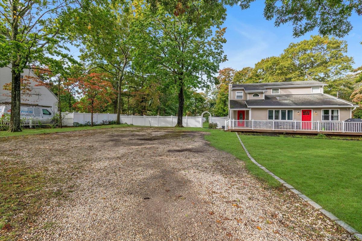 Expansive grounds featuring a wide gravel driveway, a manicured lawn with stone bordering, a wraparound deck with white railing, and a privacy fence