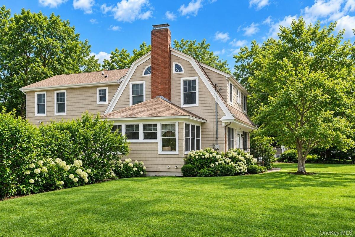 Dutch Colonial exterior featuring shingle siding, a prominent brick chimney, arched windows, and manicured landscaping