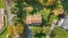 Aerial view of a residential lot featuring a shingled roof, paved driveway, expansive lawn, and mature trees