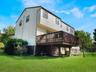 Rear exterior featuring vertical siding, a raised wood deck with balustrades, a stone retaining wall, and a large grassy lot