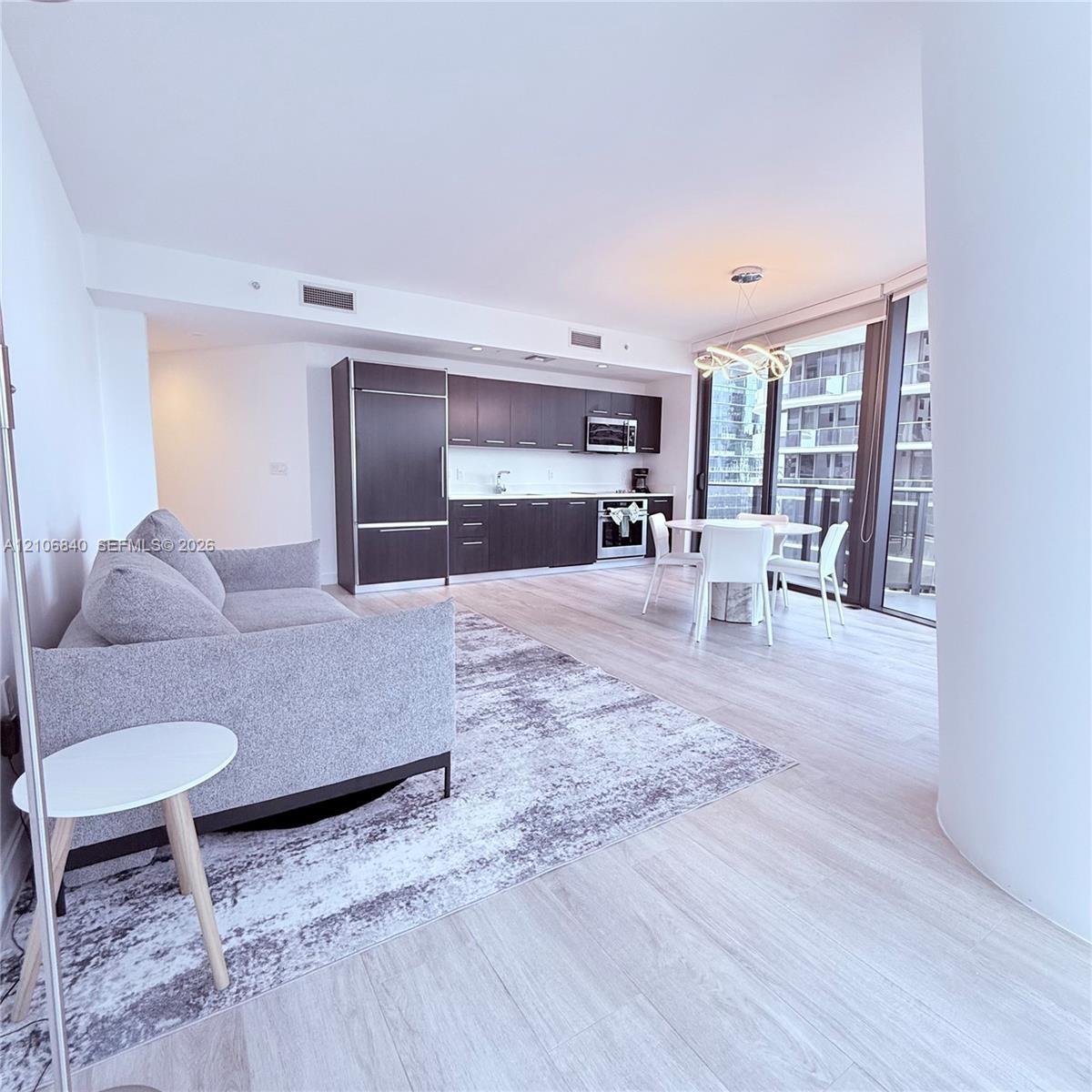 Open-concept living space featuring wood-finish flooring, dark cabinetry, stainless steel appliances, floor-to-ceiling glass, and a modern sculptural 