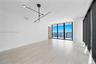 Floor-to-ceiling windows, wood-finish flooring, balcony with water backdrop, matte black geometric chandelier, and cylindrical structural column
